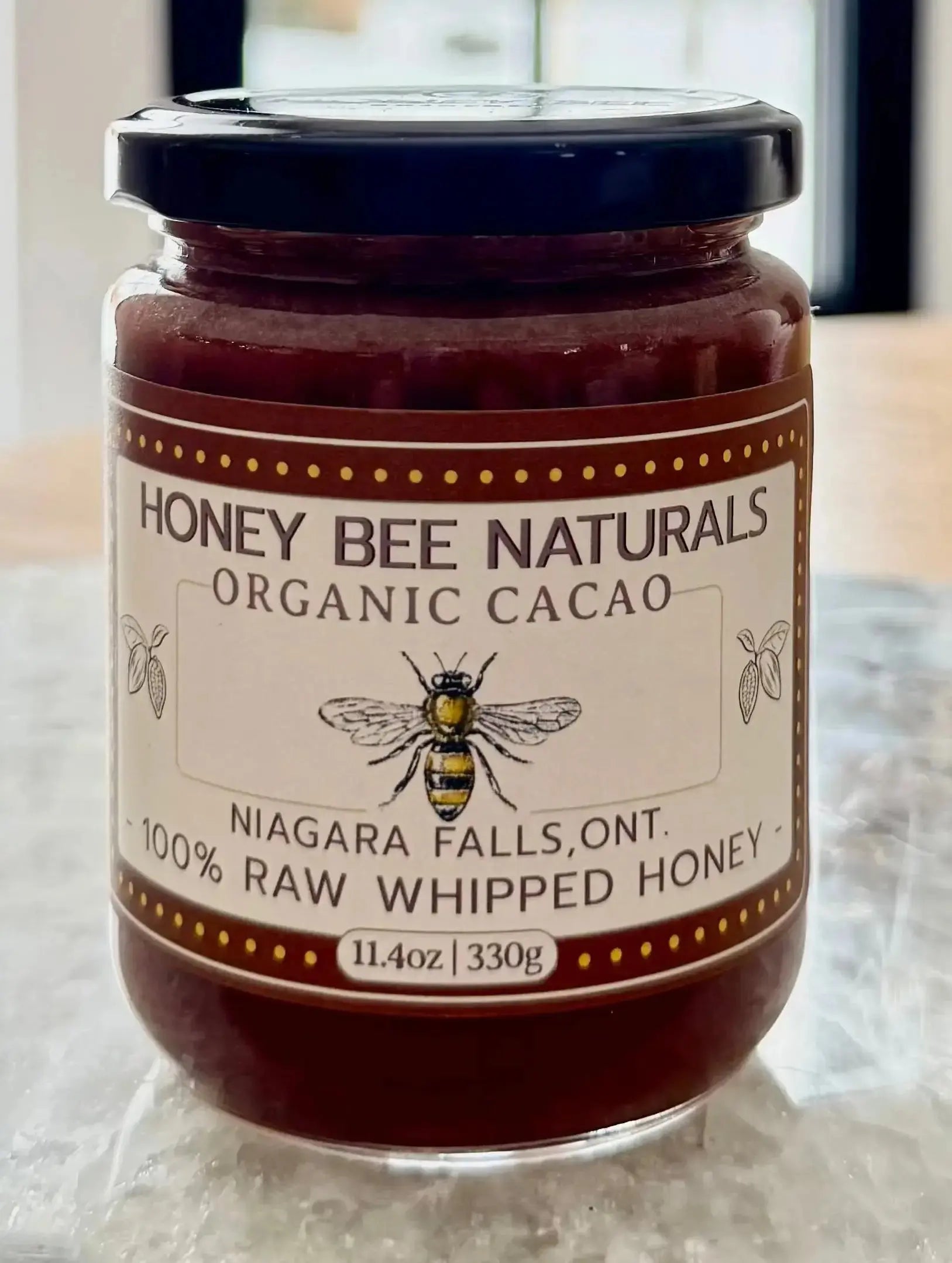 Jar of Honey Bee Naturals Organic Cacao honey on a wooden surface with a window in the background